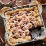 Close-up of fudgy peppermint hot chocolate brownies in a baking pan, topped with toasted mini marshmallows, melted chocolate chips, and crushed red peppermint candies. The perfect decadent holiday dessert.